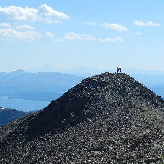 Hikers standing on the rocky top of Avalanche Peak with Yellowstone Lake in the background.