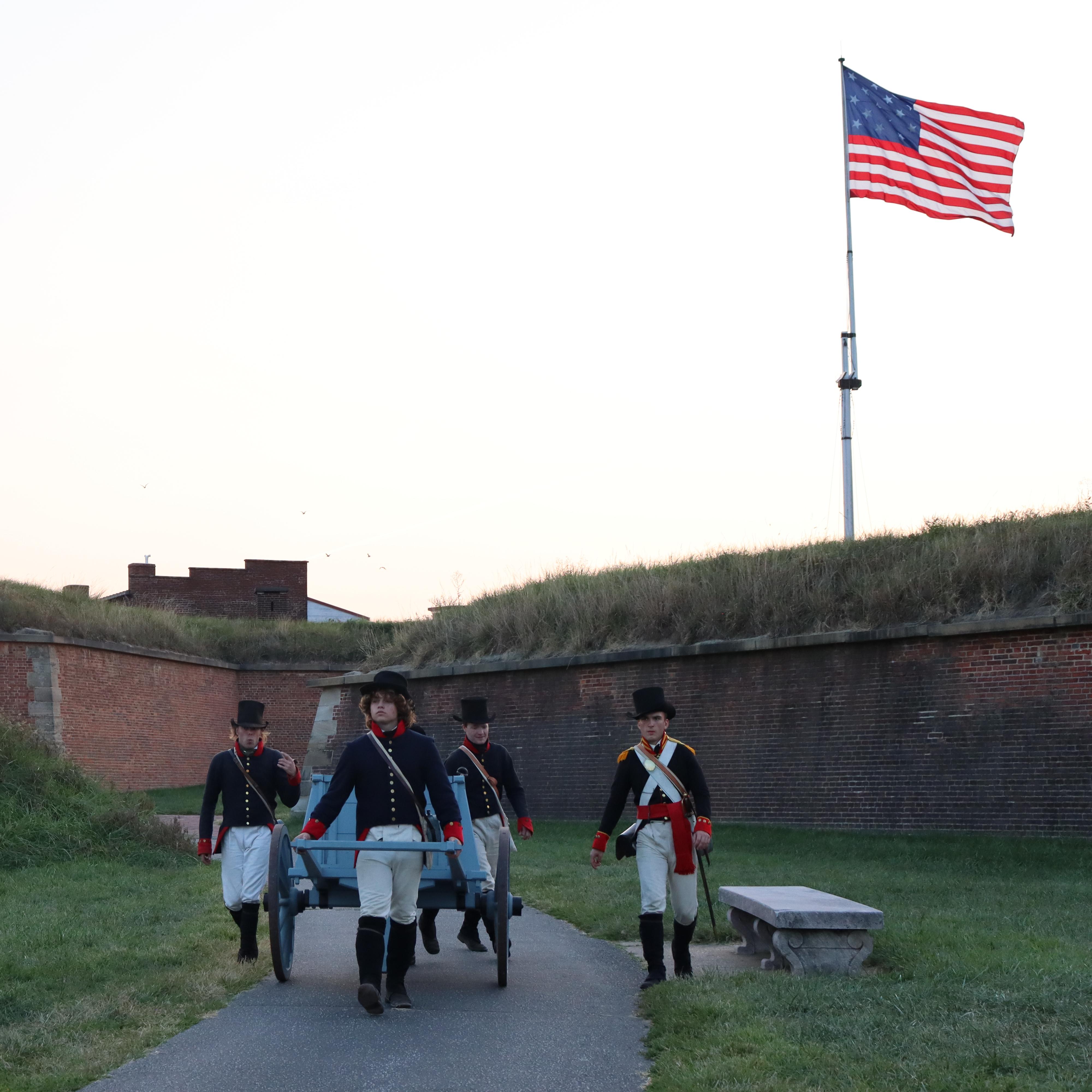 Living historians walking out of Fort McHenry, pulling a wagon, with the 17x25 flag flying