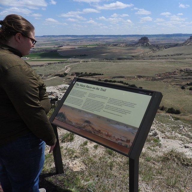 A woman reads an interpretive panel at an overlook of a valley and surrounding bluffs.