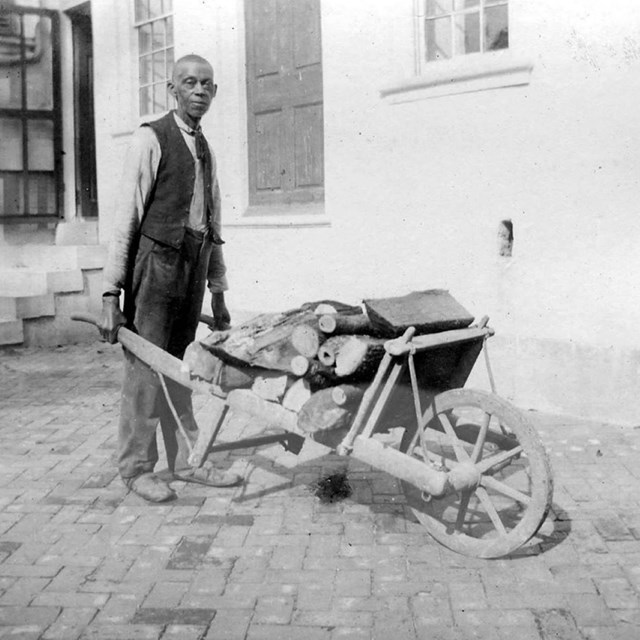 A black and white photograph of and enslaved man with wood in a wheelbarrow. 