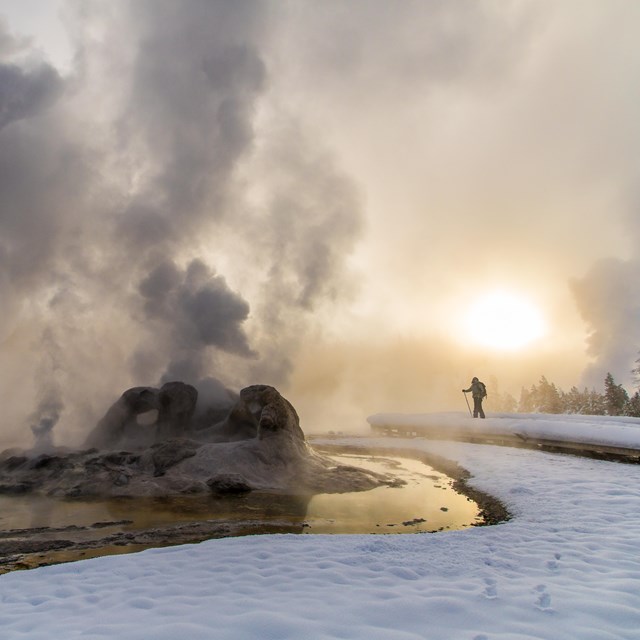 A lone visitor enjoys castle geyser from a snowy boardwalk just before sunset.