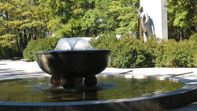 Large round marble fountain in foreground and statue of Theodore Roosevelt in background.