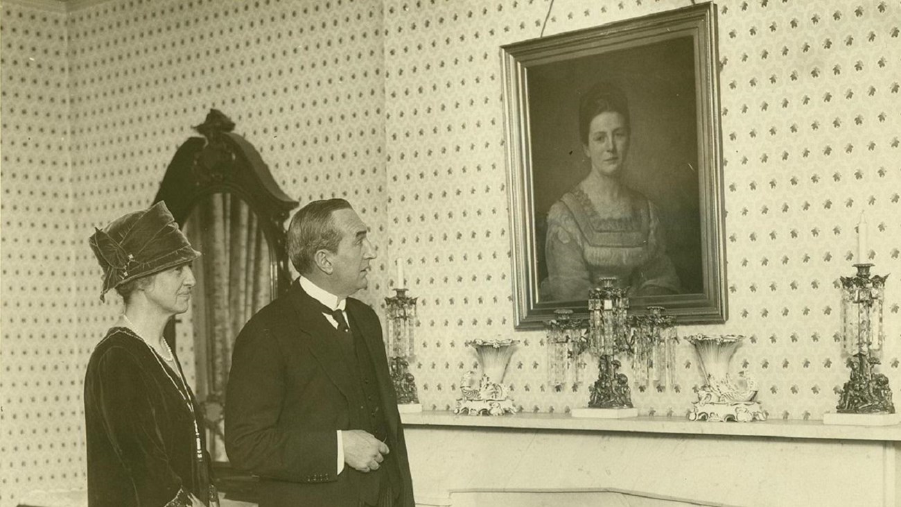 A sepia photograph of a man and a woman looking at a portrait on the wall. 