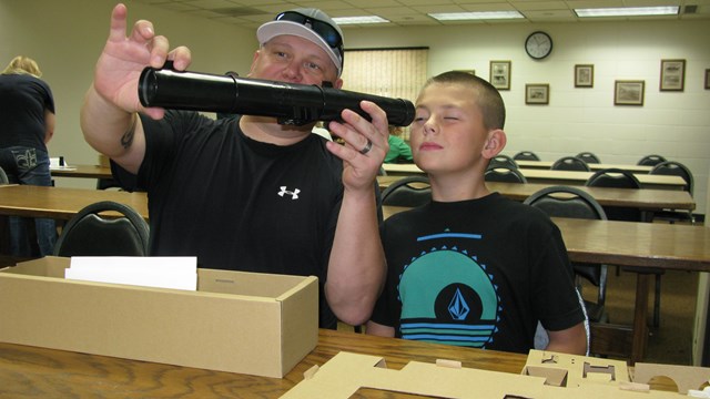 Father and son at a table peering through a telescope.