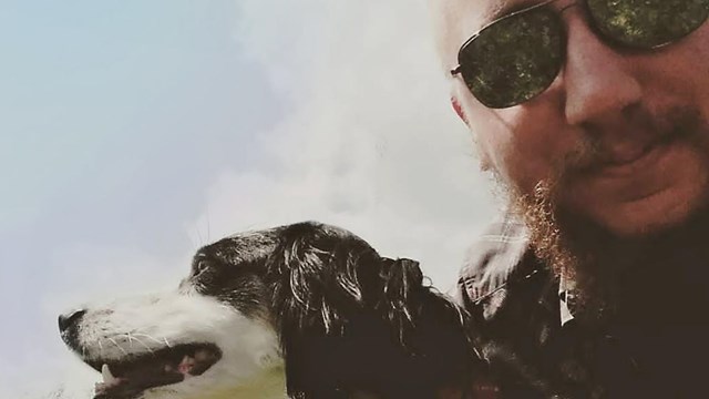 A dog wearing a harness and leash and his owner wearing sunglasses pose for a selfie.