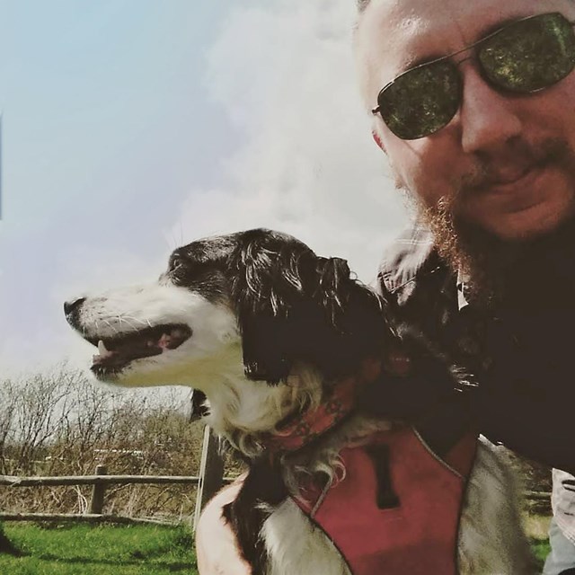 A dog wearing a harness and leash and his human wearing sunglasses take a selfie together.