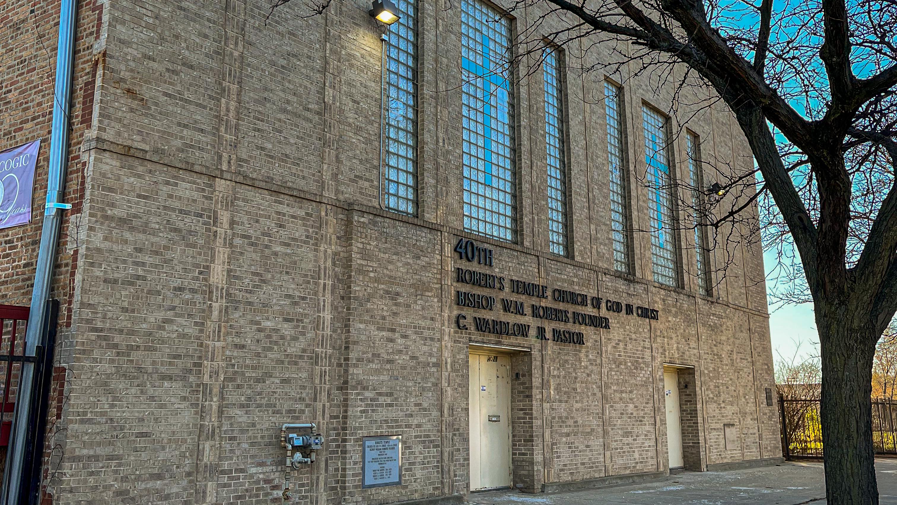 Front corner of a multi-story tan brick church with a tree in front at dusk. 