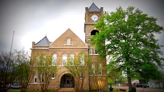A two-story brick courthouse with clock tower and tree in front.