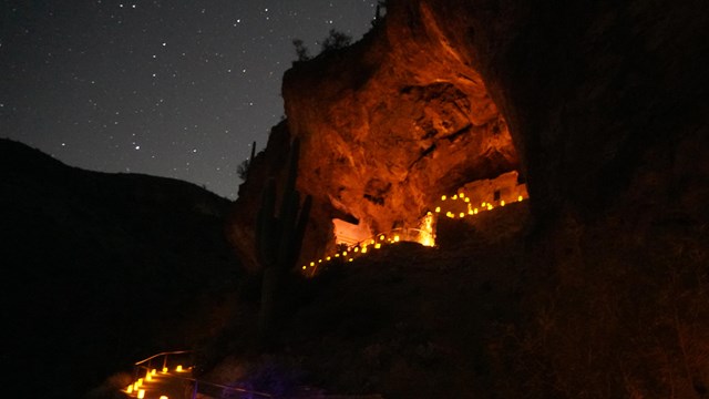 A cliff dwelling with the night sky behind it. Luminary lights line a trail and the dwelling.