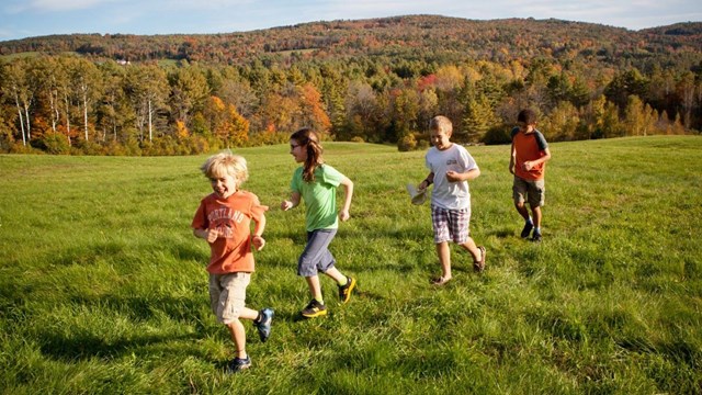 four kids run down a grassy trail with a fall forest in the background