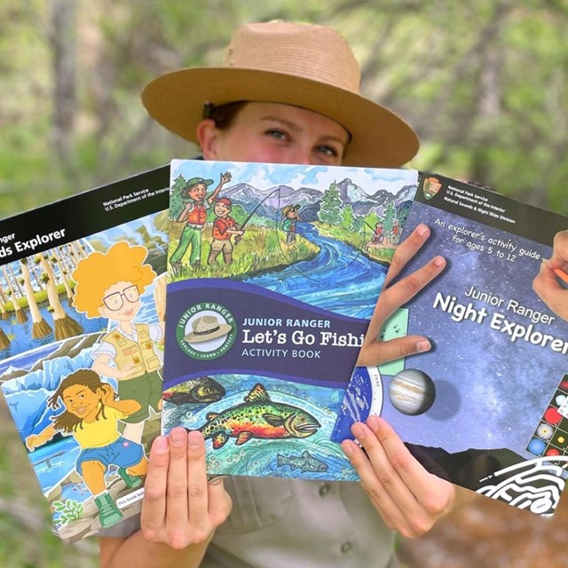 a park ranger holds up three colorful junior ranger booklets