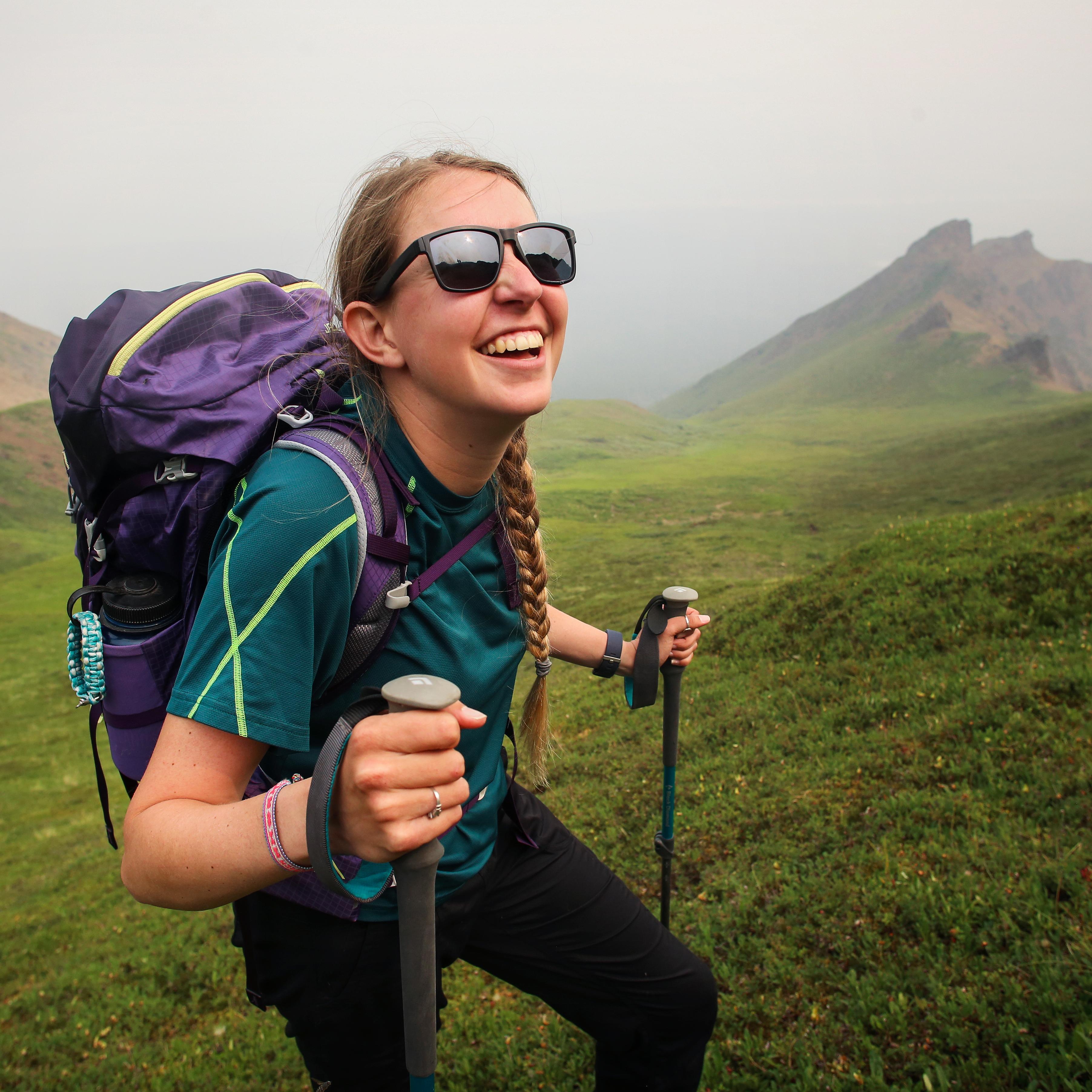 a smiling woman with sunglasses, backpack, and hiking poles