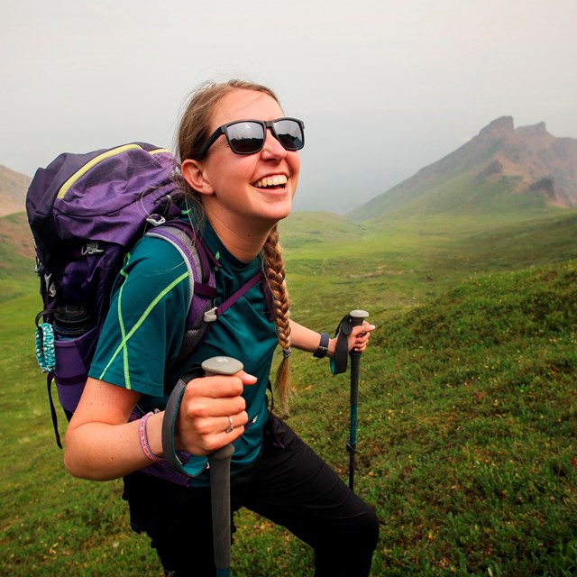 a smiling woman with sunglasses, backpack, and hiking poles