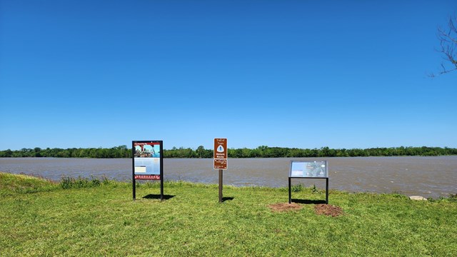 Three upright signs stand in front of a flat, broad river.