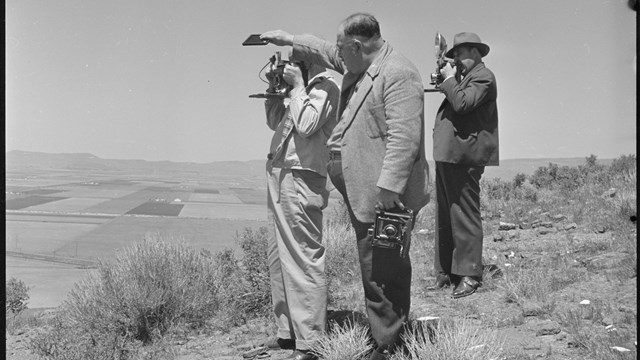 Three men stand on a hill, two pointing 1940s era cameras and one shading a camera lens