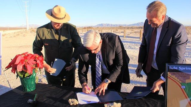 A park ranger and a man in a suit look at another man in a suit sign a document on a table