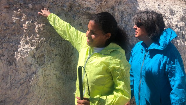 A blind woman with a cane feels a wall of sediment. She is standing next to another woman.