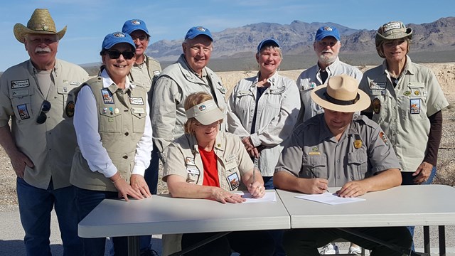 A park ranger signs a document on a table while a group of volunteers smile and look on