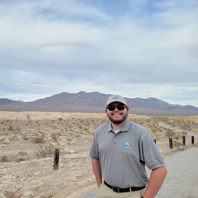 A man in a gray polo and hat poses in front of desert badlands and a mountain range.