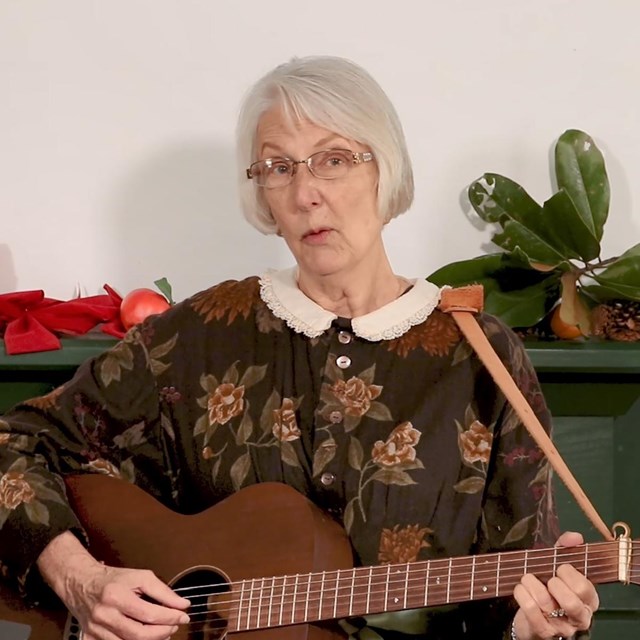 Woman wearing a hoop dress plays guitar in front of a decorated fireplace mantle. 