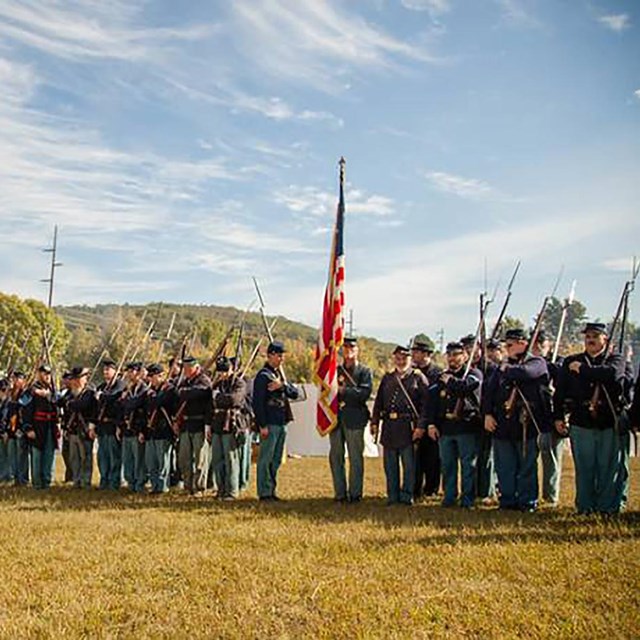 A long line of men dressed as Civil War Union soldiers. White A-frame tents visible in background.