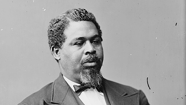 Photograph of Black man sitting at three quarter profile in a suit.