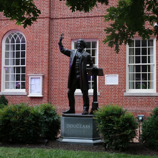 Photograph of a statue of Frederick Douglass standing in grass in front of brick buildings.