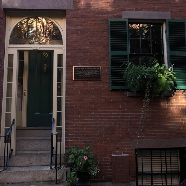 A photograph of a brick home with a deep green door & shutters, with cream door & window trimmings.
