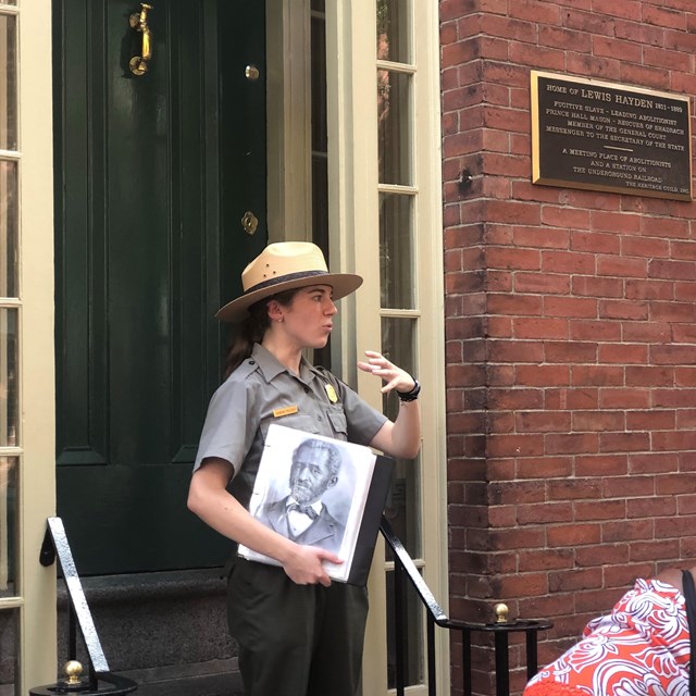 Ranger giving talk in front of a brick home. Ranger holds sketch of African American man.