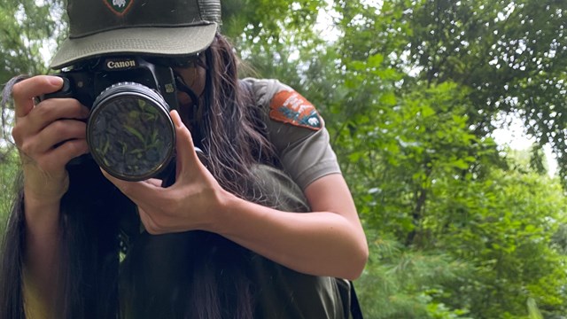 Park ranger crouches, pointing camera lens at camera, with orange newt in foreground