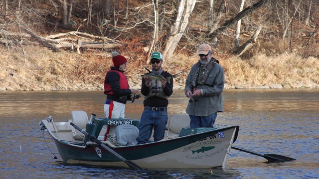 three people standing in boat with motor. Man in middle holds up large, green-gray fish.