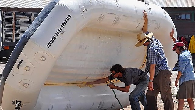 two people hold an inflatable raft up on its side while another person sprays interior with water