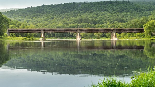 View of the roebling bridge over river against lush green hills & blue sky. Water is like a mirror.