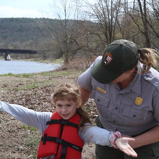Young child spreading arms out and wearing a life jacket. Ranger bends down to check life jacket.