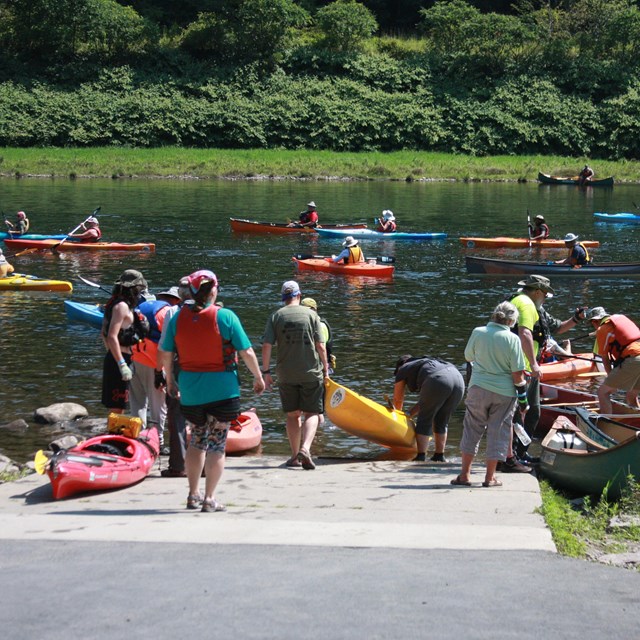 boat access ramp to river. Pairs of people in T-shirts and shorts carry kayaks and canoes to water.