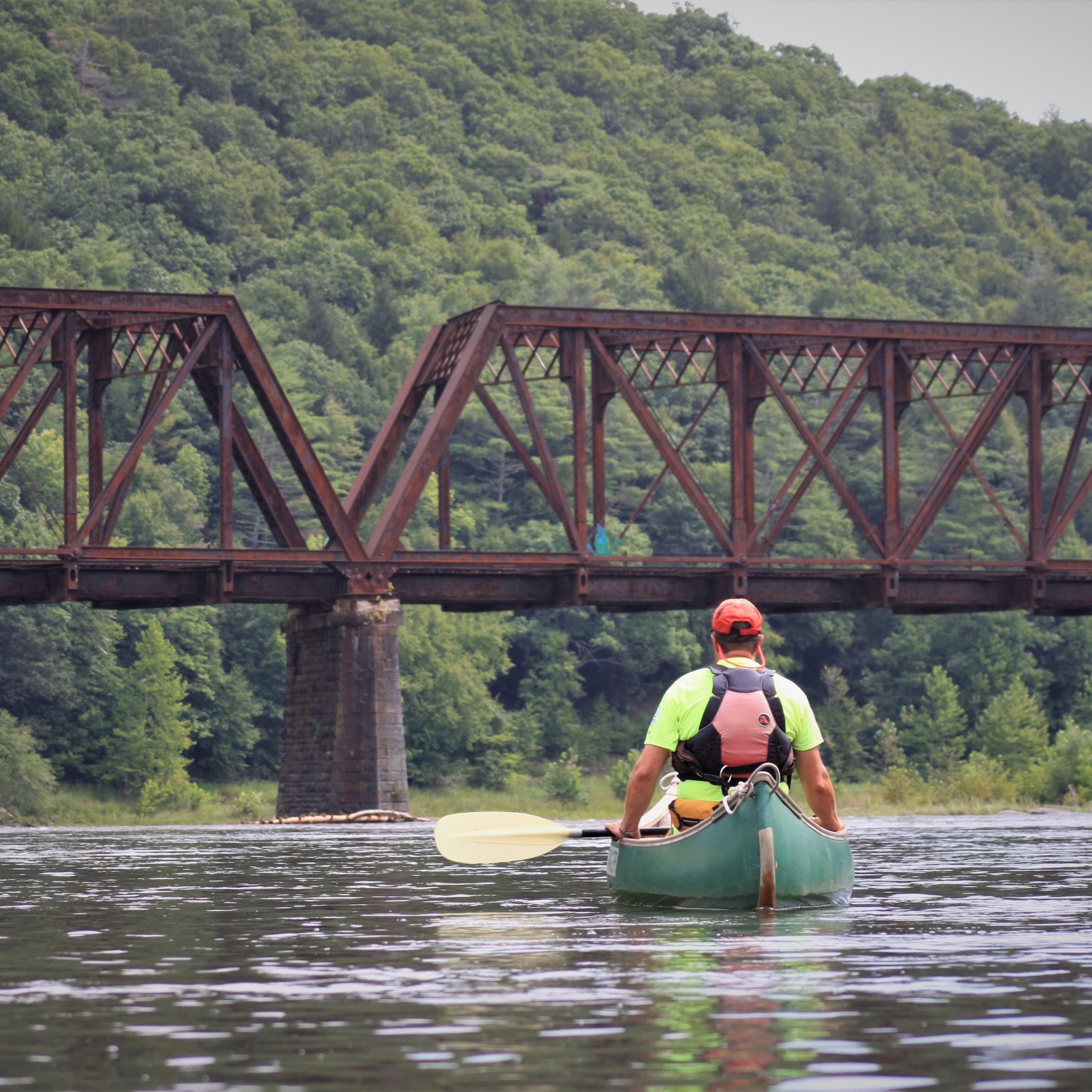 Man in life jacket sitting in canoe on the river with a bridge and forested hills in background.