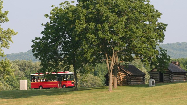 A red and yellow trolley travels up a wooded hill past log huts.