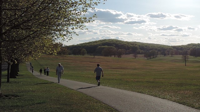 Walkers enjoy a late afternoon stroll along a paved path through a large grassy field.