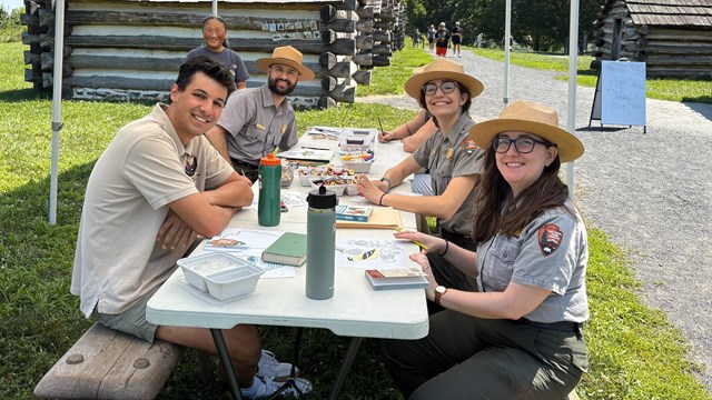 smiling park rangers sit at a table with a smiling intern near log huts