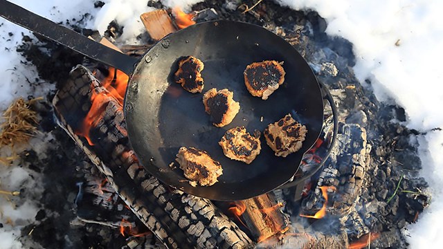 balls of dough cook in a metal skillet placed on top of an open wood fire