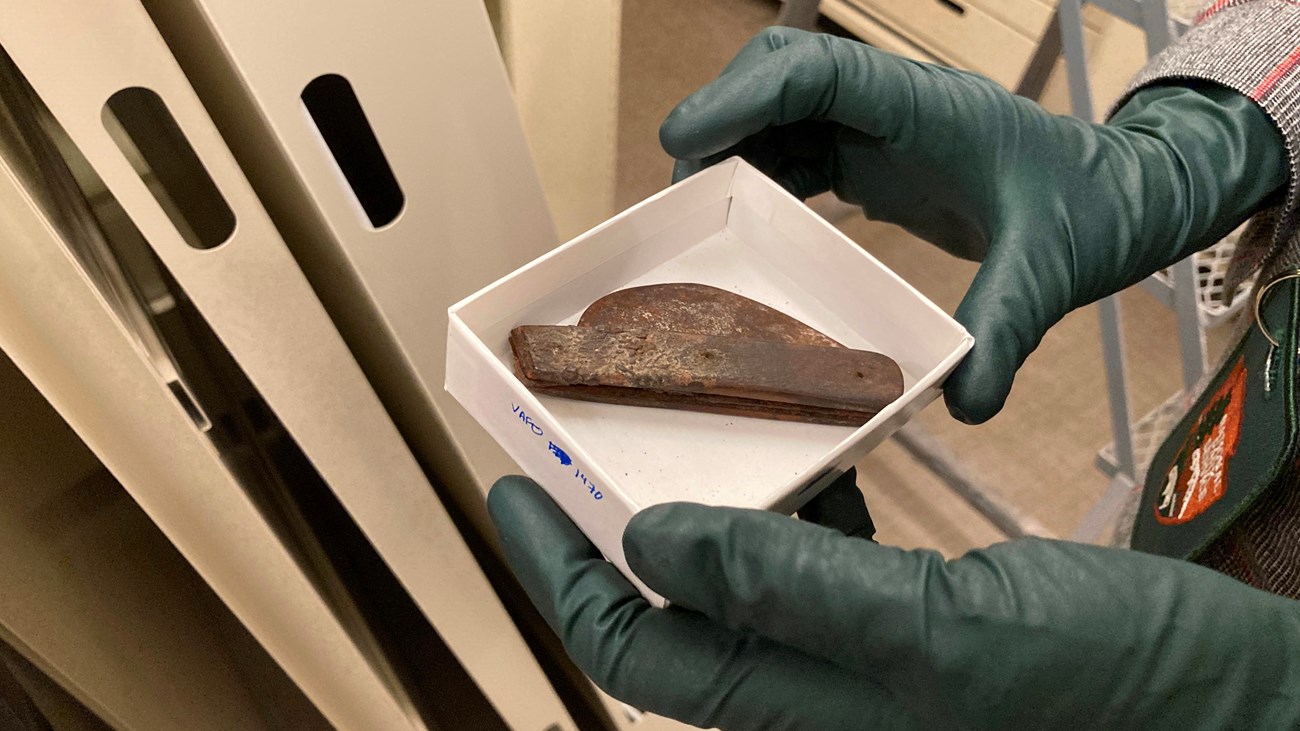 A small box held by gloved hands contains a small folding knife artifact.