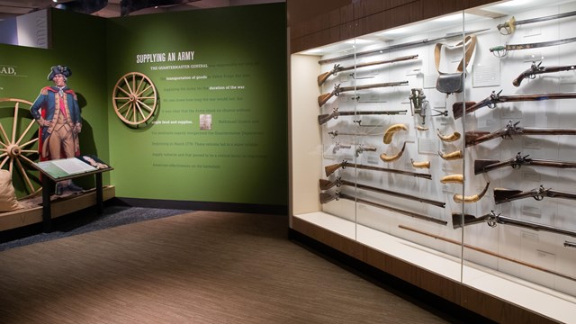 Firearms and edged weapons mounted in an exhibit case next to an exhibit wall with text.