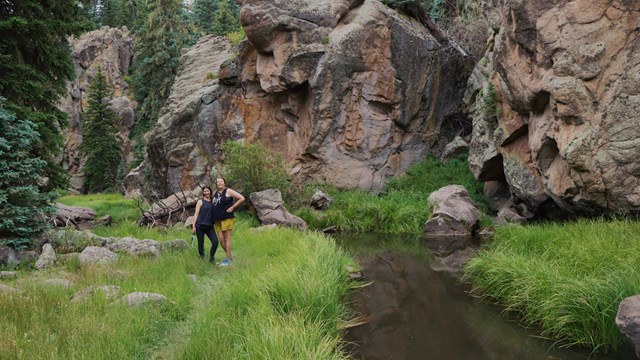 Two women smile while standing next to a narrow stream with big boulders along the bank.