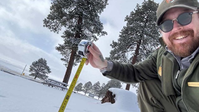 A park ranger kneels in the snow with a tape measure. Snow reaches the 6-inch mark.