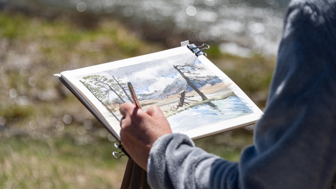 A closeup of an artist's hands as they paint a mountain scene in a notebook.