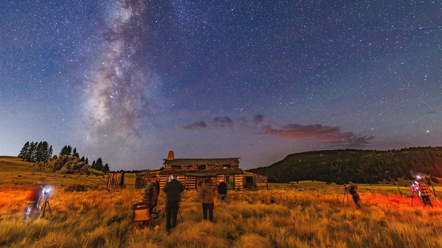 Several photographers scattered across a grassy field with a cabin and the starry night sky overhead