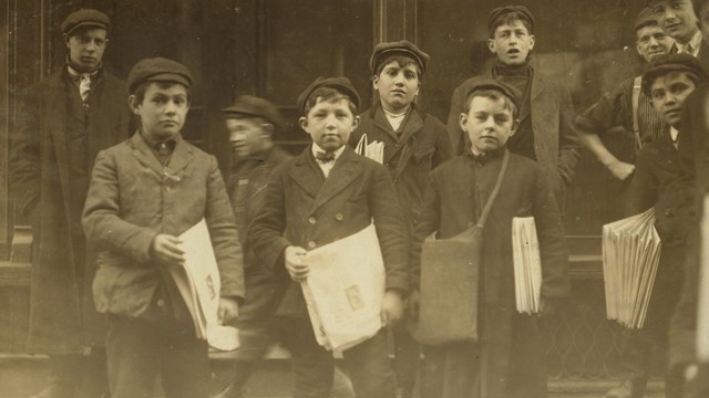 A group of young boys wearing knickers and selling newspapers.