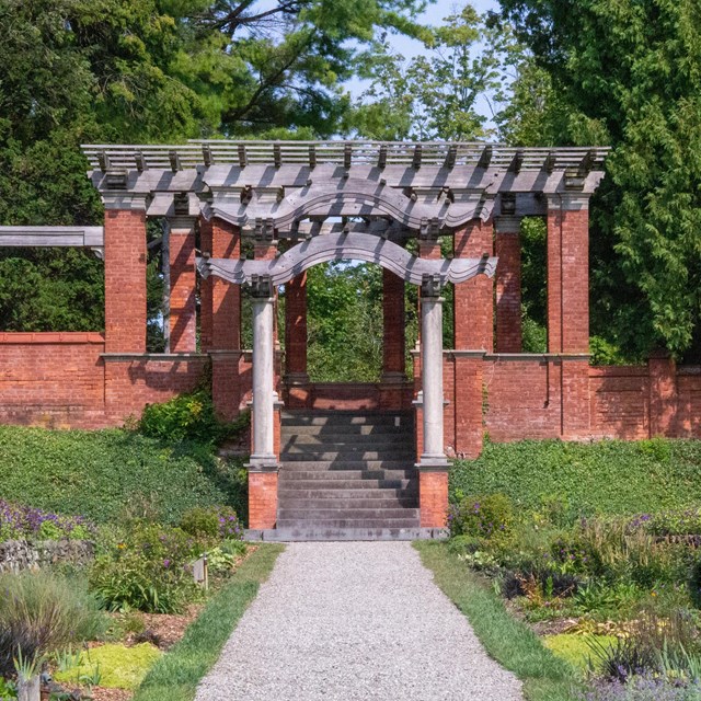 A gravel path bordered by flowers leading to a red brick and timber pergola.