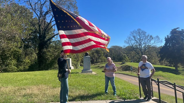 Man dressed in historic uniform carrying a replica flag talking to visitors