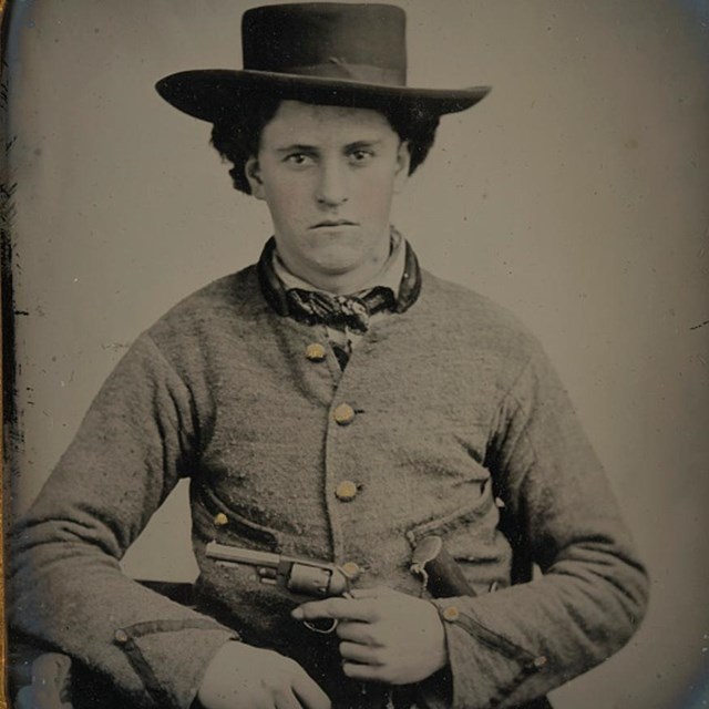 A Confederate soldier in gray uniform holding a pistol wearing a slouch hat.